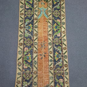 Könnte beinhalten: Rechteckiger Teppich mit einem orangefarbenen Mittelstreifen und floralen Mustern in Blau, Grün und Beige. Der Teppich hat ein traditionelles Design mit aufwendigen Details und einem strukturierten Aussehen. Die Kanten sind mit einem dezenten Rand versehen.