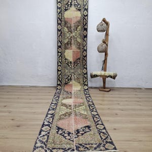 Peut inclure: Un long tapis rectangulaire avec un motif central de formes géométriques roses, beiges et marron alternées. Le tapis a une bordure bleu foncé avec des motifs complexes. Le tapis est exposé sur un sol en bois.