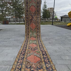 May include: An antique runner rug with a detailed floral pattern. The rug features a central design in shades of coral and green, framed by a navy blue border with yellow accents. The rug is displayed outdoors on a paved surface.