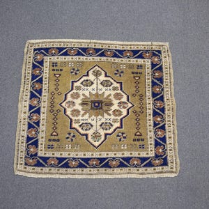 May include: Square rug with an intricate geometric pattern. The rug features a central design in cream and brown, surrounded by a border of blue and brown floral motifs. The rug has a beige fringe.