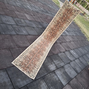 Pode incluir: Um tapete longo e retangular com um intrincado padrão floral em tons de castanho, bege e verde. O tapete é exibido ao ar livre, mostrando seu design e textura. As bordas do tapete são finalizadas com uma borda detalhada.