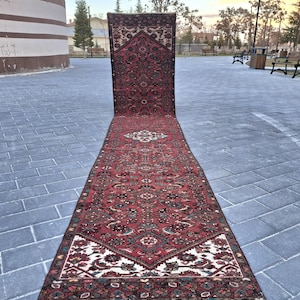 May include: A long, rectangular, red rug with an intricate, repeating pattern. The rug features a central medallion design and borders with floral and geometric motifs. The rug is displayed outdoors on a brick surface.