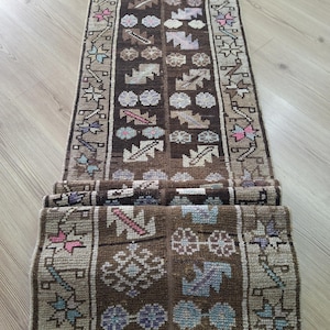Puede incluir: Una alfombra de pasillo larga y estrecha con un campo central de color marrón oscuro decorado con motivos geométricos y florales repetitivos en tonos rosa, azul claro y blanco. El borde muestra un patrón de estrellas repetitivo sobre fondo beige.