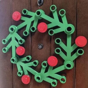 May include: A green and red Lego wreath hanging on a brown door. The wreath is constructed of green Lego pieces with red Lego circles evenly spaced around the wreath. The wreath is circular and has a festive appearance.