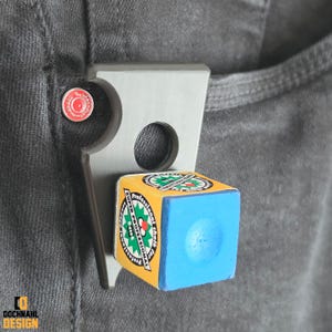 Könnte beinhalten: Ein Metallclip mit rundem Loch und einem kleinen roten und weißen kreisförmigen Objekt, das an einer Tasche befestigt ist. Ein blauer Kreidewürfel mit gelben Seiten wird von dem Clip gehalten. Auf dem Kreidewürfel steht "Super Professional Chalk".