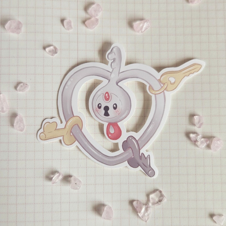 May include: A white and gray key-shaped charm with a heart-shaped outline. The key has a small, round, gray head with a keyhole and a red heart in the center. The charm is attached to a silver chain.