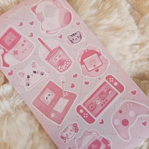 May include: Pink and white sticker sheet with a variety of cute gamer-themed illustrations, including a cat wearing headphones, a game console, a boba tea, and a cat holding a game controller. The sheet also includes hearts, stars, and other decorative elements. The text "Sluggy Atelier" is in the bottom right corner.
