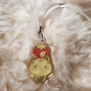 May include: A clear acrylic charm depicting a cartoon apple character topped with a red strawberry. It has a silver-coloured clasp and a white cord. The design has a cute, whimsical aesthetic, suitable for a bag or keyring.