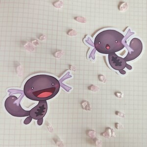 May include: Two purple axolotl stickers with white outlines and pink accents. The axolotls are smiling with their mouths open and have small, black eyes. They are both in a cartoon style.