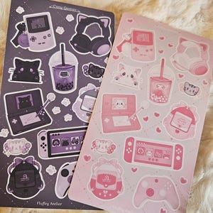 May include: Two sheets of stickers featuring a pink and a gray color scheme. The stickers feature various cute illustrations of cats, game consoles, and other gaming-related items. The text "Cozy Gamer" and "Fluffy Atelier" is printed on the sheets.