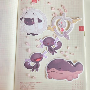 May include: Five colorful cartoon stickers featuring different Pokemon characters. The stickers are on a white gridded notebook page with pink glitter scattered around them. The characters include a white sheep with black markings, a purple and black axolotl, a purple and black axolotl with a wide smile, a purple and pink whale, and a silver key with a heart-shaped loop.