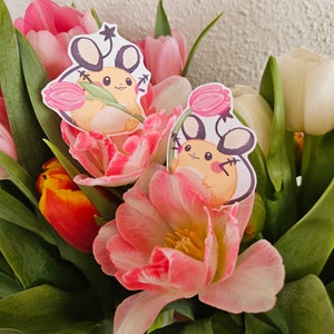 May include: Two pink and white tulips with cartoon stickers of a small, yellow, mouse-like creature holding a pink flower. The stickers are on the tulips.