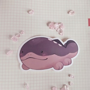 May include: A sticker of a cartoon purple and pink creature with a wide, flat body and small legs. The creature has a friendly expression and is lying on its belly.