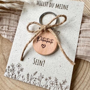 Op de afbeelding: Een houten sleutelhanger met het woord "Patentante" en een gegraveerd hart, bevestigd aan een kaart met de tekst "Willst du meine sein?" en bloemenillustraties. De sleutelhanger is vastgebonden met een touwstrik.