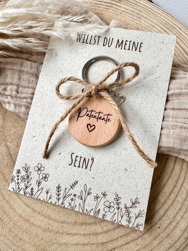 Op de afbeelding: Een houten sleutelhanger met het woord "Patentante" en een gegraveerd hart, bevestigd aan een kaart met de tekst "Willst du meine sein?" en bloemenillustraties. De sleutelhanger is vastgebonden met een touwstrik.
