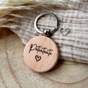 Könnte beinhalten: Ein Holzschlüsselanhänger mit einem silbernen Ring und einem herzförmigen Anhänger. Der runde Holzanhänger trägt die Aufschrift "Patentante" und ein eingraviertes Herzsymbol. Der Schlüsselanhänger wird auf einem neutralfarbenen Stoff präsentiert.