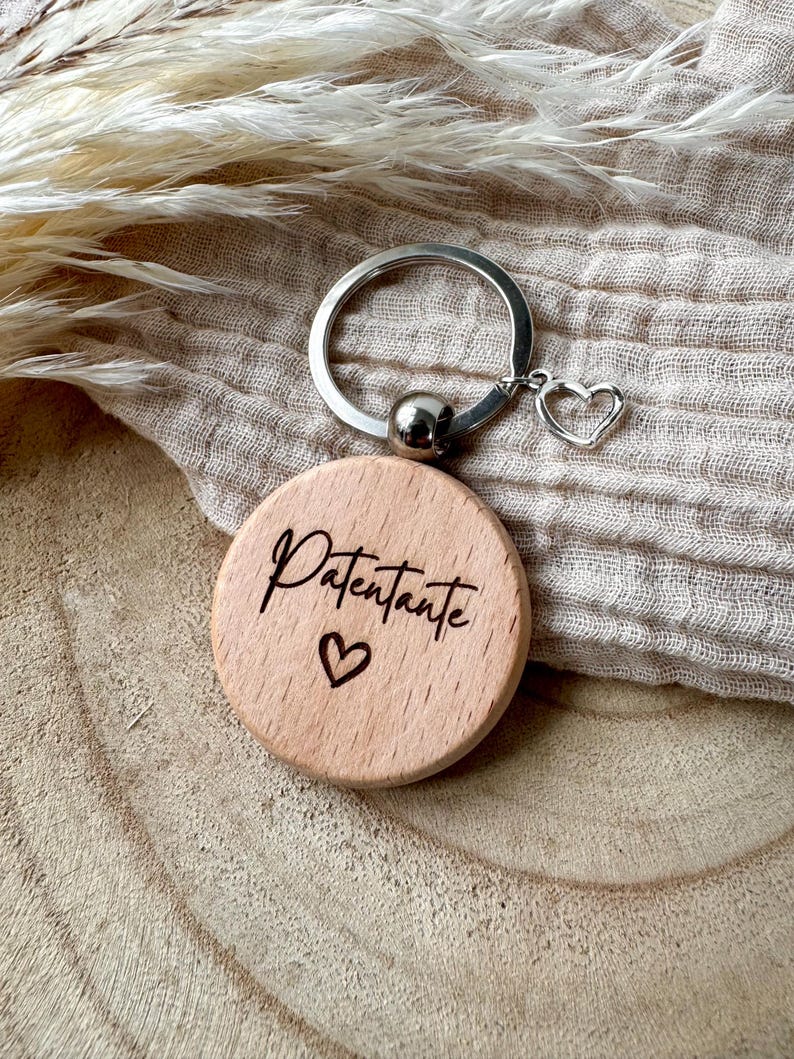 Op de afbeelding: Een houten sleutelhanger met een zilveren ring en een hartvormige bedel. De ronde houten hanger heeft de tekst "Patentante" en een gegraveerd hartsymbool. De sleutelhanger wordt gepresenteerd op een neutraal gekleurde stof.