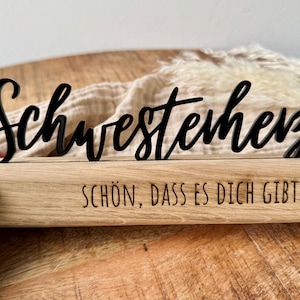 Könnte beinhalten: Ein Holzschild mit den Worten "Schwesterherz" in schwarzer Schrift und dem Text "Schön, dass es dich gibt" mit einem kleinen Herzsymbol.