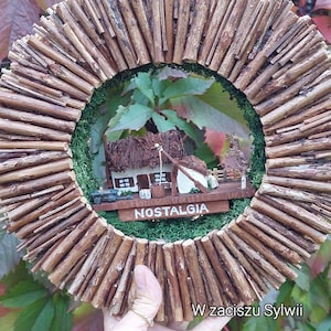 Może przedstawiać: Drewniana wieniec z miniaturowym domkiem w środku. Domek jest drewniany i ma strzechowy dach. Wieniec jest ozdobiony zielonym mchem i napisami "NOSTALGIA" i "W zaciszu Sylwii".