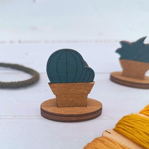 以下が含まれることがあります： 茶色とティール色の木製サボテンと多肉植物の切り抜き。サボテンは縦線が入った丸い形で、多肉植物は尖った葉をしています。植物は丸い台座に置かれています。黄色の糸と緑色のロープも表示されています。
