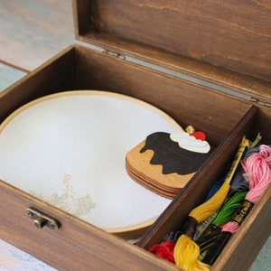 May include: A wooden box with a hinged lid, containing a white embroidery hoop, a wooden cake charm, and a selection of colorful embroidery floss.