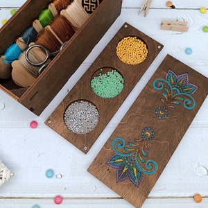 May include: A wooden box with three compartments for storing beads. The compartments are filled with yellow, green, and white beads. The box is decorated with a floral design.
