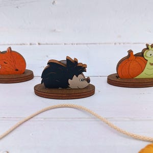May include: Three wooden animal figurines on a white wooden surface. The figurines are a fox, a hedgehog, and a snail carrying a pumpkin. The figurines are all painted in bright colours.