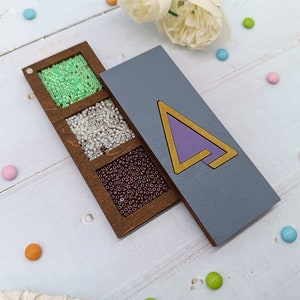 May include: A wooden box with three compartments filled with different coloured beads. The box is next to a purple and gold wooden triangle.