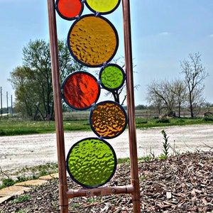 May include: A copper pipe garden sculpture with colorful stained glass circles in shades of red, orange, yellow, and green. The sculpture is standing on a bed of wood chips.