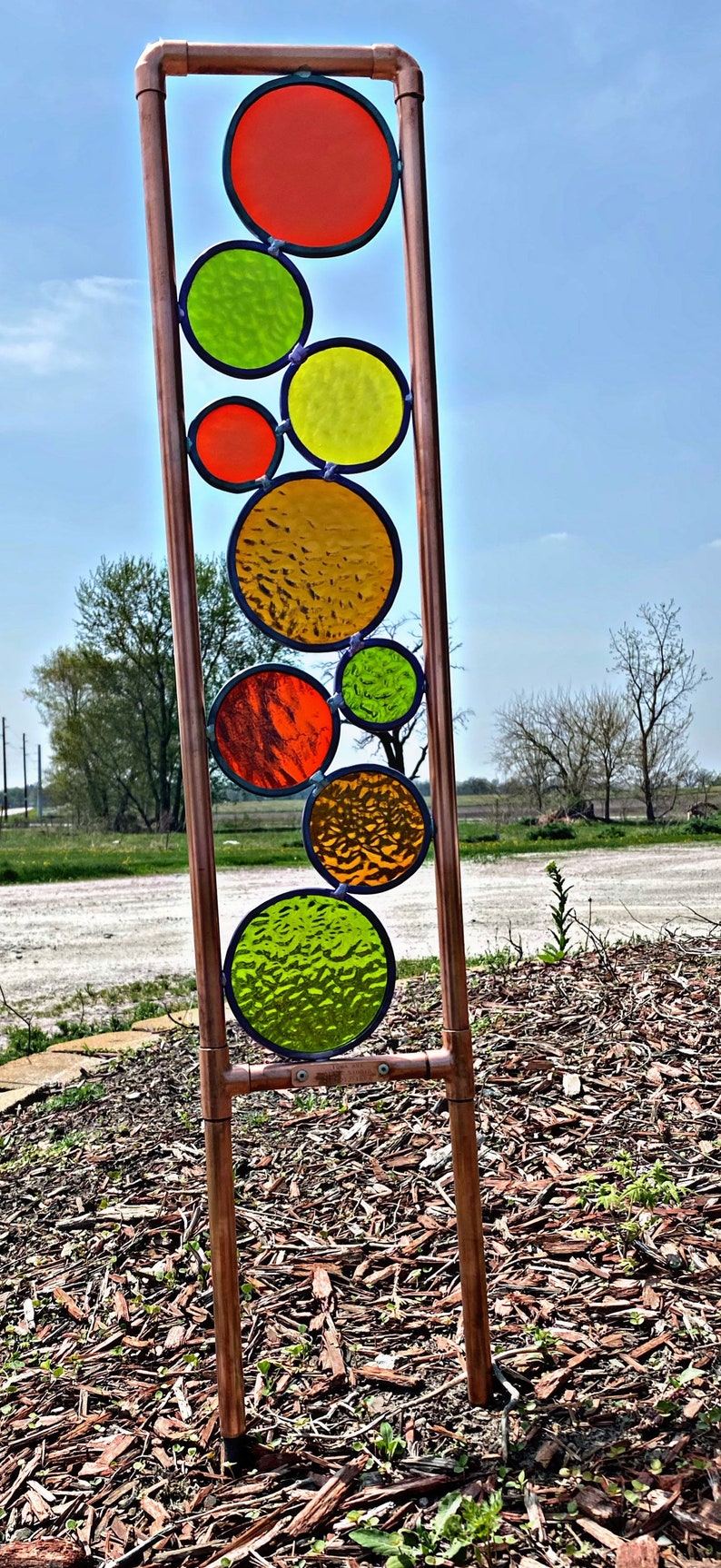 May include: A copper pipe garden sculpture with colorful stained glass circles in shades of red, orange, yellow, and green. The sculpture is standing on a bed of wood chips.