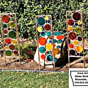 May include: Three colorful stained glass panels with a circular design, each panel is mounted on a copper stand. The panels are made of various colors of glass, including red, orange, yellow, green, and blue. The panels are arranged in a row, with the copper stands visible in the foreground. The panels are set against a backdrop of green foliage.