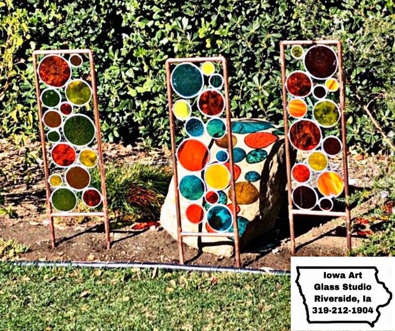 May include: Three colorful stained glass panels with a circular design, each panel is mounted on a copper stand. The panels are made of various colors of glass, including red, orange, yellow, green, and blue. The panels are arranged in a row, with the copper stands visible in the foreground. The panels are set against a backdrop of green foliage.