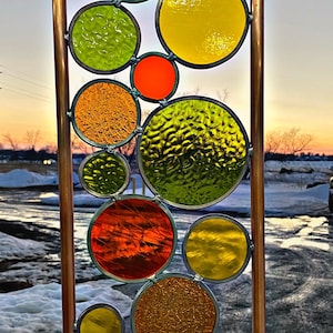 May include: A copper pipe frame holds a stained glass panel with colorful circles in shades of red, orange, yellow, and green. The circles are arranged in a random pattern and create a modern and abstract design. The panel is set against a snowy background with a sunset in the distance. The business card for Iowa Art Glass Studio is visible in the bottom right corner.