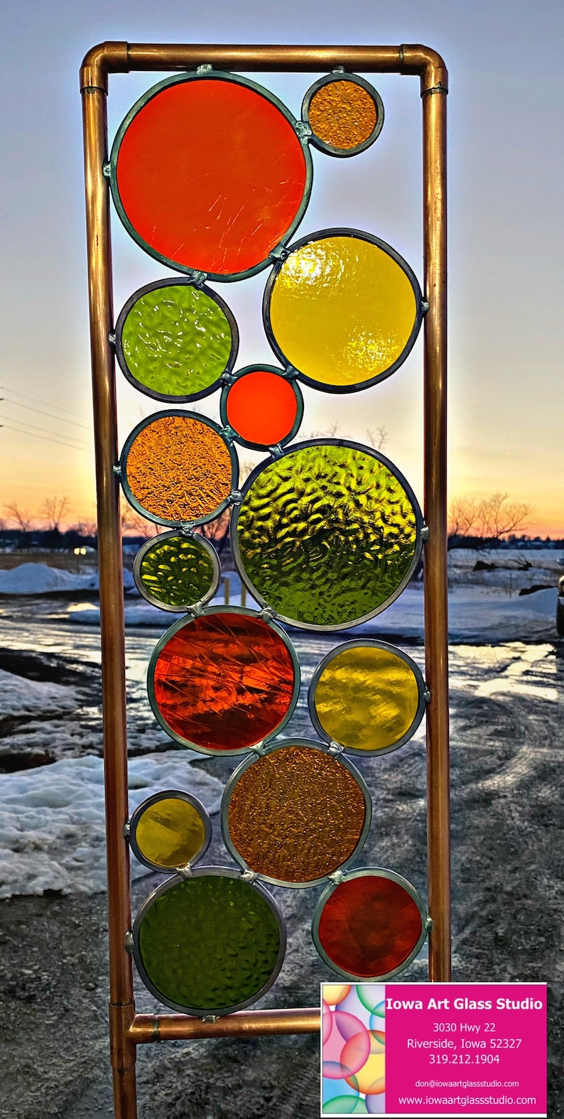 May include: A copper pipe frame holds a stained glass panel with colorful circles in shades of red, orange, yellow, and green. The circles are arranged in a random pattern and create a modern and abstract design. The panel is set against a snowy background with a sunset in the distance. The business card for Iowa Art Glass Studio is visible in the bottom right corner.