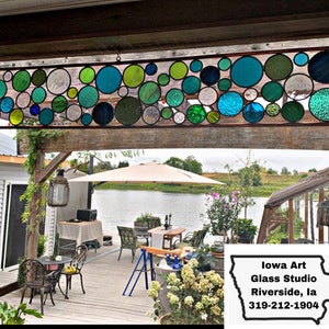 Puede incluir: Un panel de vitral con un dise&ntilde;o de c&iacute;rculos coloridos en tonos de verde, azul y blanco. La ventana est&aacute; colgada en un marco de madera y se ve desde un patio con un lago en el fondo. El texto "Iowa Art Glass Studio Riverside, IA 319-212-1904" es visible en la esquina inferior derecha de la imagen.