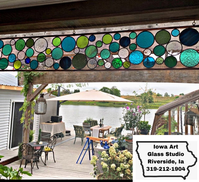 Puede incluir: Un panel de vitral con un dise&ntilde;o de c&iacute;rculos coloridos en tonos de verde, azul y blanco. La ventana est&aacute; colgada en un marco de madera y se ve desde un patio con un lago en el fondo. El texto "Iowa Art Glass Studio Riverside, IA 319-212-1904" es visible en la esquina inferior derecha de la imagen.