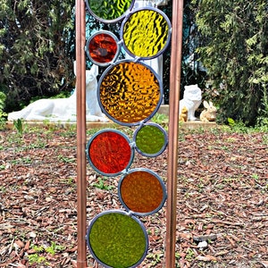 May include: A copper pipe garden sculpture with colorful stained glass circles in shades of red, orange, yellow, and green. The sculpture is standing in a bed of brown wood chips.