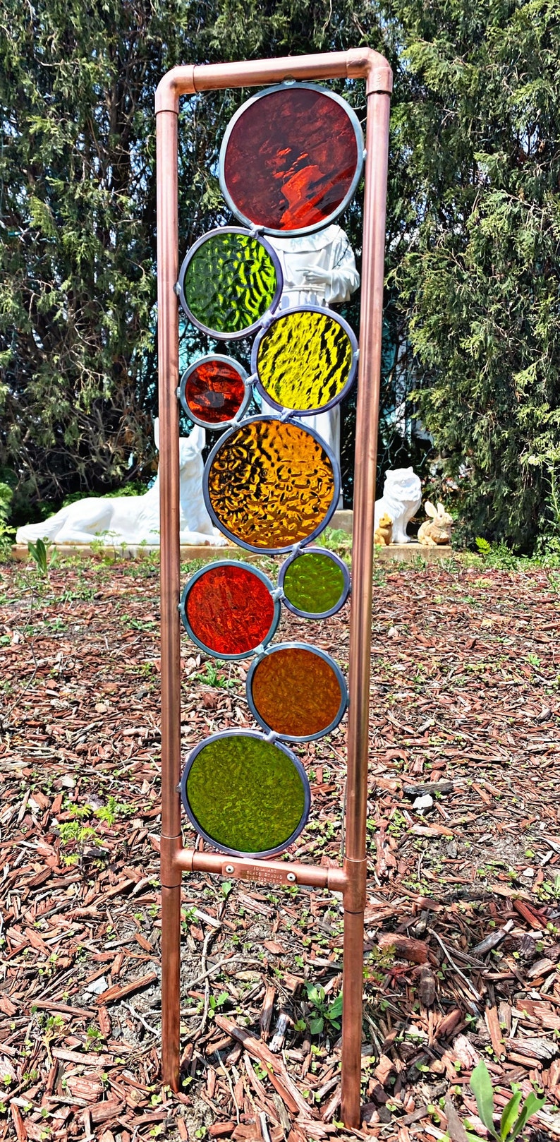 May include: A copper pipe garden sculpture with colorful stained glass circles in shades of red, orange, yellow, and green. The sculpture is standing in a bed of brown wood chips.