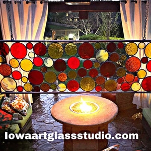 May include: A stained glass window panel with a circular design featuring red, yellow, and brown glass circles. The panel is hanging from a chain and is in front of a patio with a fire pit.