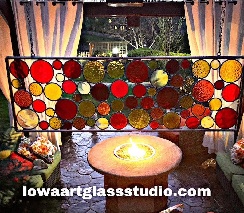 May include: A stained glass window panel with a circular design featuring red, yellow, and brown glass circles. The panel is hanging from a chain and is in front of a patio with a fire pit.