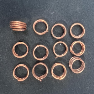 May include: A collection of 14 copper jump rings, including one larger ring with multiple coils. The rings are all circular and have a smooth, shiny finish.