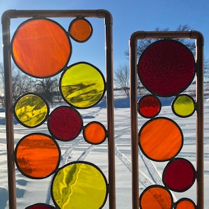 May include: Two copper pipe framed stained glass panels with colorful circles in shades of red, orange, and yellow. The panels are set against a snowy background.