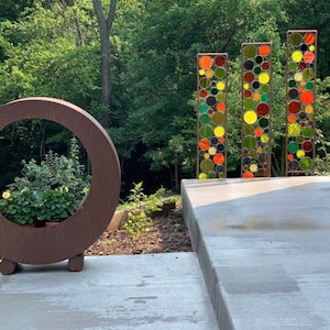 May include: A circular metal planter with a brown finish and a large opening in the center. The planter is sitting on a concrete patio with a backdrop of green trees. Three tall metal screens with colorful circular glass pieces are visible in the background.