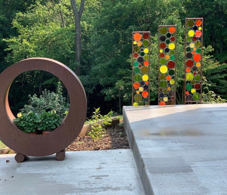 May include: A circular metal planter with a brown finish and a large opening in the center. The planter is sitting on a concrete patio with a backdrop of green trees. Three tall metal screens with colorful circular glass pieces are visible in the background.