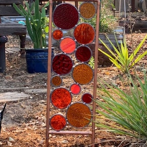 May include: A copper pipe garden sculpture with stained glass circles in shades of red, orange, and brown.