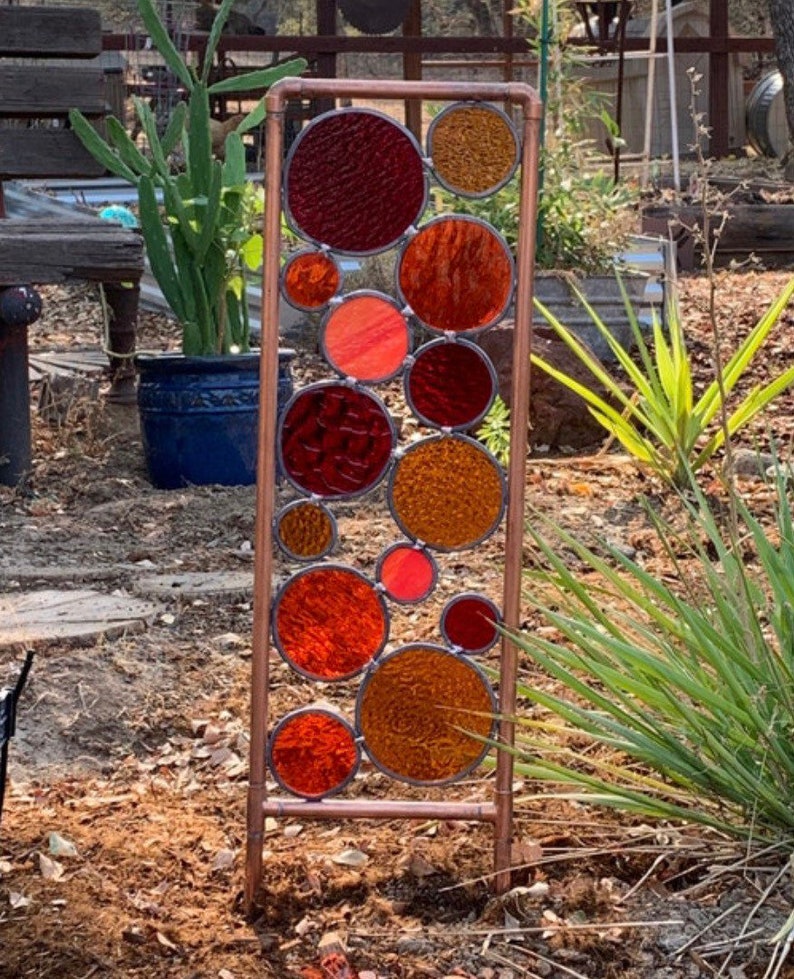 May include: A copper pipe garden sculpture with stained glass circles in shades of red, orange, and brown.