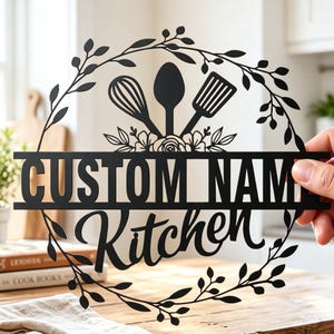 Op de afbeelding: Een rond wandbord van zwart metaal met een bladvormige kransrand. In het midden bevinden zich een garde, een lepel en een spatel boven de tekst "CUSTOM NAME" en het woord "Kitchen" in een sierlijk lettertype, alles binnen het ontwerp.
