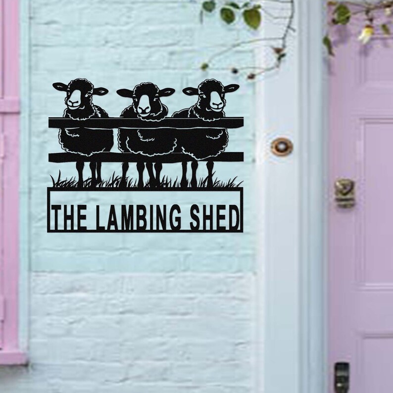 May include: Black metal sign featuring three sheep perched on a fence, with the words "THE LAMBING SHED" below. The sign is mounted on a light blue brick wall, with a pink door and trim visible.