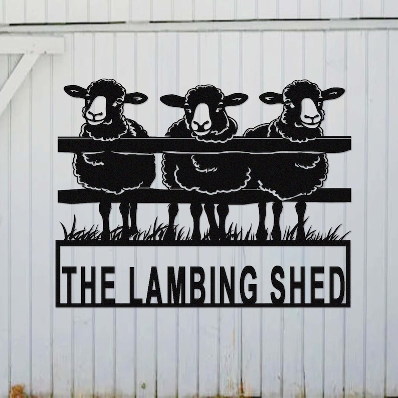 May include: Black metal sign featuring three sheep peering over a fence, with the words "THE LAMBING SHED" in a rectangular frame below. The sign is mounted on a white wooden background.