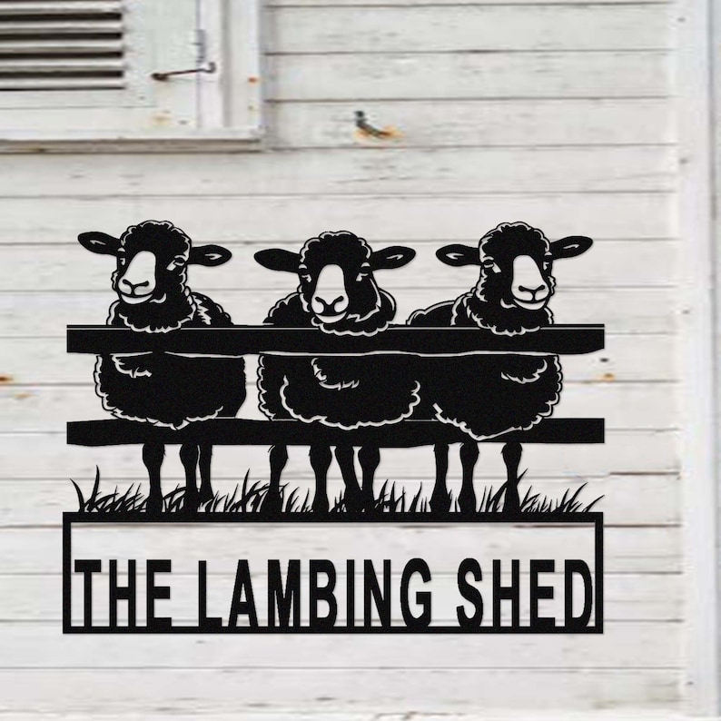 May include: Black metal sign featuring three sheep peering over a fence. The sign includes the text "THE LAMBING SHED" in a rectangular frame, with grass at the base. The sheep have fluffy wool and are set against a white background.
